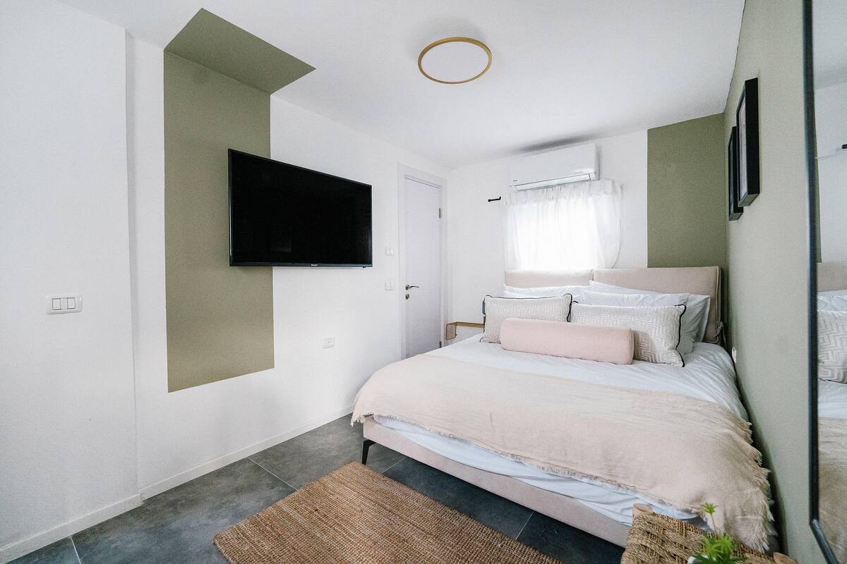 Une chambre à coucher avec un lit, un téléviseur à écran plat fixé au mur, une fenêtre avec des rideaux et un petit tapis sur le sol.