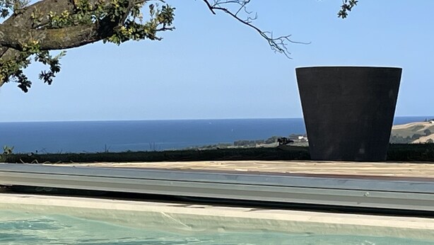 En pool med klart vatten, en stor, mörk konstruktion och utsikt över havet.