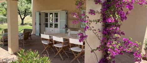 An outdoor patio with a wooden table and chairs, a covered area, and a wall with purple flowers.
