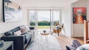 A modern living room with a sofa, a small round table, and a view through a glass door to an outdoor area. - Villa with Spa and Breathtaking Views of Dandenong Ranges (Kalorama)