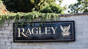 Ein Schild mit dem Namen „RAGLEY“ und einem Wappen, an einer Steinmauer befestigt, die mit Efeu überwuchert ist.