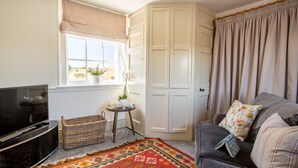 A cozy living room with a television, a wicker basket, a small table with a teapot, a sofa with floral cushions, and a large wardrobe.