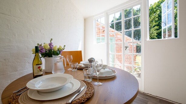 A dining table set with plates, cutlery, a pitcher, and glasses, with a window view of a brick building and greenery.