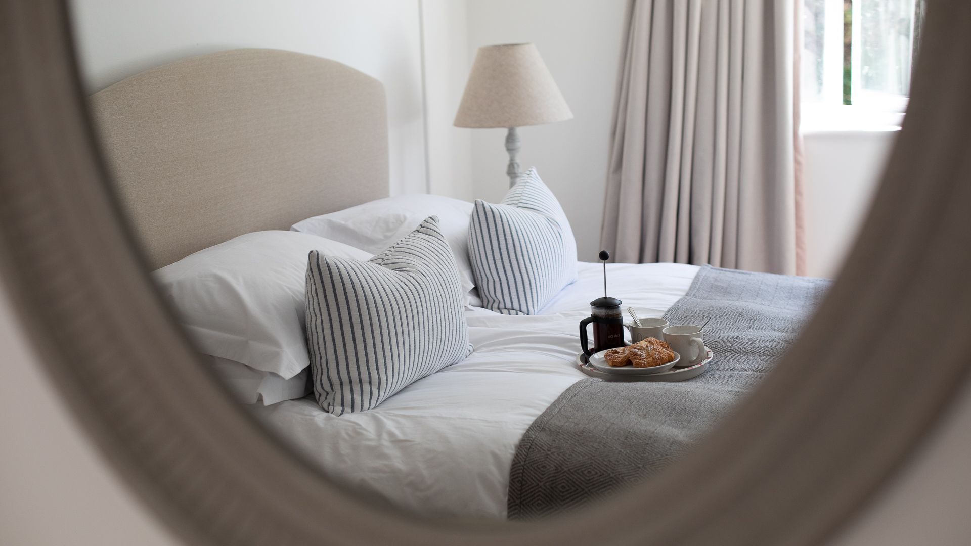 Una cama bien hecha con almohadas a rayas, una mesita de noche con una cafetera y pasteles, y una lámpara.