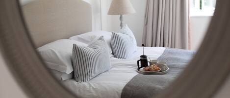 Ein ordentlich gemachtes Bett mit gestreiften Kissen, ein Nachttisch mit Kaffeekocher und Gebäck sowie eine Lampe.