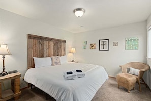 Un dormitorio con una cama grande, dos lámparas de noche, una silla de mimbre y decoración en la pared.