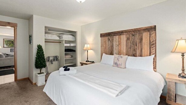 A hotel room with a large bed, a wooden headboard, a closet with shelves, a small plant, and a nightstand with a lamp. - Downtown Leavenworth | King Bed + Parking (Leavenworth)