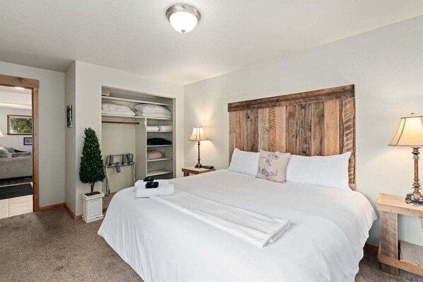 A hotel room with a large bed, a wooden headboard, a closet with shelves, a small plant, and a nightstand with a lamp. - Adorable and Affordable in Downtown Leavenworth (Leavenworth)