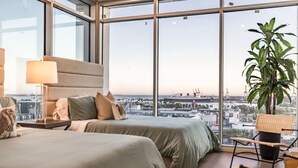 A modern hotel room with two beds, a large window offering a city view, a potted plant, and a chair.