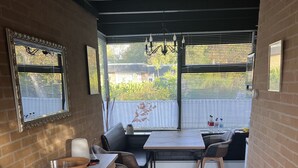 Un'area pranzo con tavolo e sedie, un mobiletto in legno, un tappeto decorato e vista attraverso una finestra con persiane.