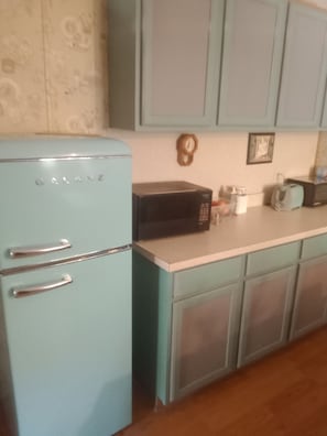 A kitchen with a light blue refrigerator, a microwave, and a toaster. There are cabinets with frosted glass doors and a wooden clock on the wall.