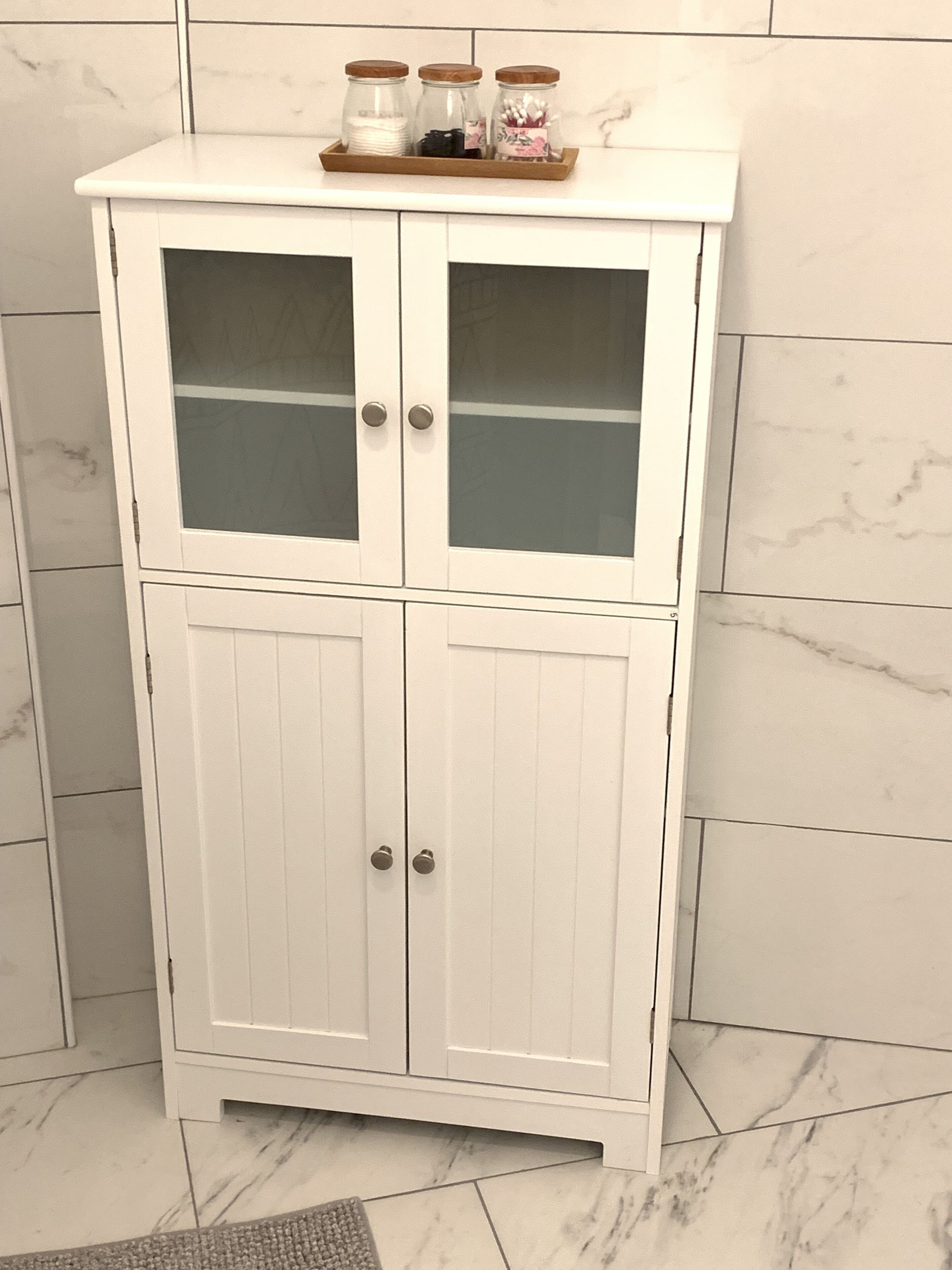 A white cabinet with two glass doors and two solid doors, topped with a wooden tray holding jars.