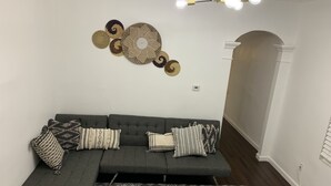 En moderne stue med en grå hjørnesofa, puder med mønster, et dekorativt vægophæng og en lysekrone.
