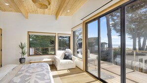 Ein modernes Schlafzimmer mit einem großen Bett, einem weißen Sessel und Blick ins Freie durch große Fenster.