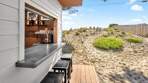 Eine Strandbar mit einer Holzterrasse und Sitzgelegenheiten im Freien, mit Blick auf eine sandige Landschaft und vereinzelter Vegetation.