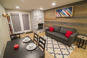 A cozy living area with a stone fireplace, a gray sofa with red cushions, a dining table set for two, and a bookshelf with books. - Cozy Main St 1BR/Next to Ski Access & FREE Shuttle (Breckenridge)