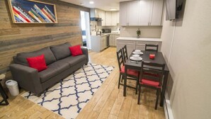 Een knus woongedeelte met een grijze bank, rode kussens, een eettafel met stoelen, een keuken en een wandgemonteerde televisie.