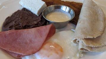 A plate with a fried egg, ham, beans, a piece of fried meat, a piece of bread, and a small bowl of sauce.