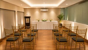Une salle de conférence avec des rangées de chaises, un pupitre, une table agrémentée de compositions florales et un appareil de climatisation.