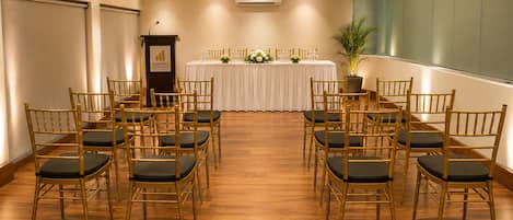 A conference room with rows of chairs, a podium, a table with floral arrangements, and an air conditioning unit.