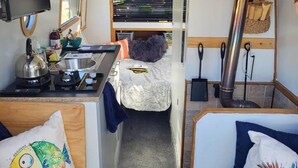 A compact RV interior with a kitchenette, a bed, and a seating area with two blue sofas. - Charming Baby Narrowboat Apsley Marina (Hemel Hempstead)