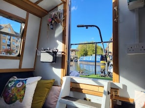 Kapean jokilaivan sisätilat: siellä on sänky, pieni pöytä ja ikkuna, josta avautuu näkymä kanavalle, jossa on veneitä ja rakennuksia.
