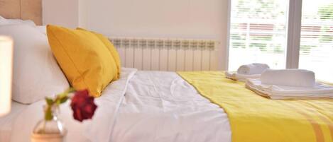 A neatly made bed with white and yellow bedding, a vase with a red flower, and a window with blinds.