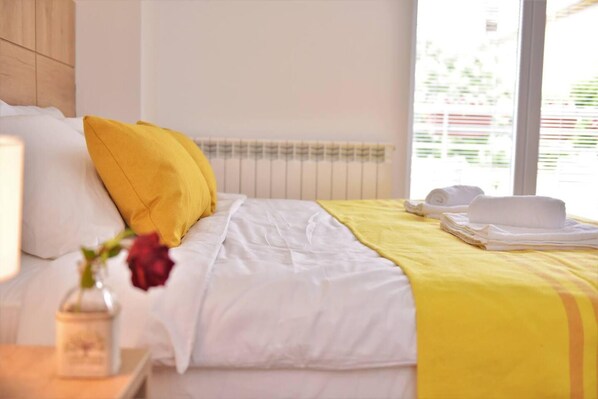 Ein ordentlich bezogenes Bett mit weißer und gelber Bettwäsche, eine Vase mit einer roten Blume und ein Fenster mit Jalousien.