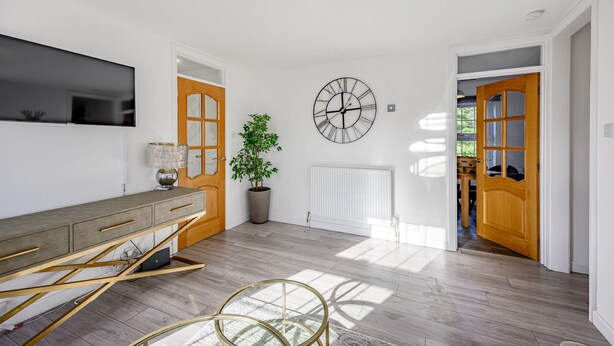 A modern living room with a wooden floor, a grey sofa, a TV mounted on the wall, a large clock, and a potted plant. - Carlisle - Apartment - Sleeps 5 - Pet-friendly (Carlisle)