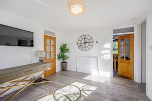 A modern living room with a wooden floor, a grey sofa, a TV mounted on the wall, a large clock, and a potted plant. - Carlisle - Apartment - Sleeps 5 - Pet-friendly (Carlisle)