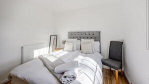 A neatly made bed with a grey upholstered headboard, a grey chair, and a white radiator. - Carlisle - Apartment - Sleeps 5 - Pet-friendly (Carlisle)