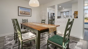 Un comedor con una mesa de madera, sillas tapizadas en verde, un alfombra estampado y una lámpara colgante.