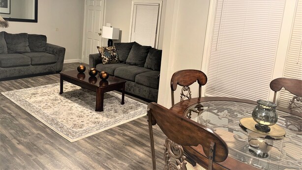 A living room with a sofa, coffee table, and chairs. There is a door, a window with blinds, and a lamp.