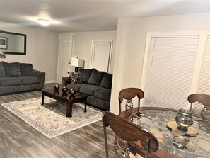 A living room with a sofa, coffee table, and chairs. There is a door, a window with blinds, and a lamp. - The Hideaway - Executive/Vacation Rental Home (Jonesboro)