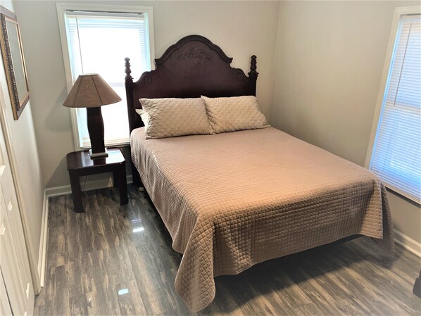 A bedroom with a wooden bed, a bedside table with a lamp, and a window with blinds. - The Hideaway - Executive/Vacation Rental Home (Jonesboro)