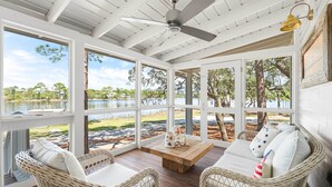 A covered outdoor seating area with wicker furniture, a wooden table, and large windows overlooking a natural landscape.