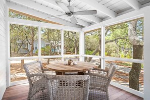 A covered patio with wicker furniture and a ceiling fan, large windows offering a view of trees and a house.