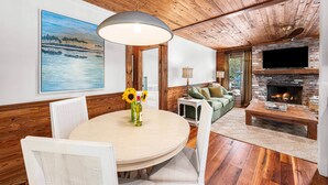 A cozy living room with a wooden ceiling, a round dining table with chairs, a sofa, a TV mounted on a brick wall, and a fireplace.