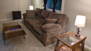 A living room with a brown sofa, a glass coffee table, a wooden side table with a lamp, a TV mounted on the wall, and a window with curtains.