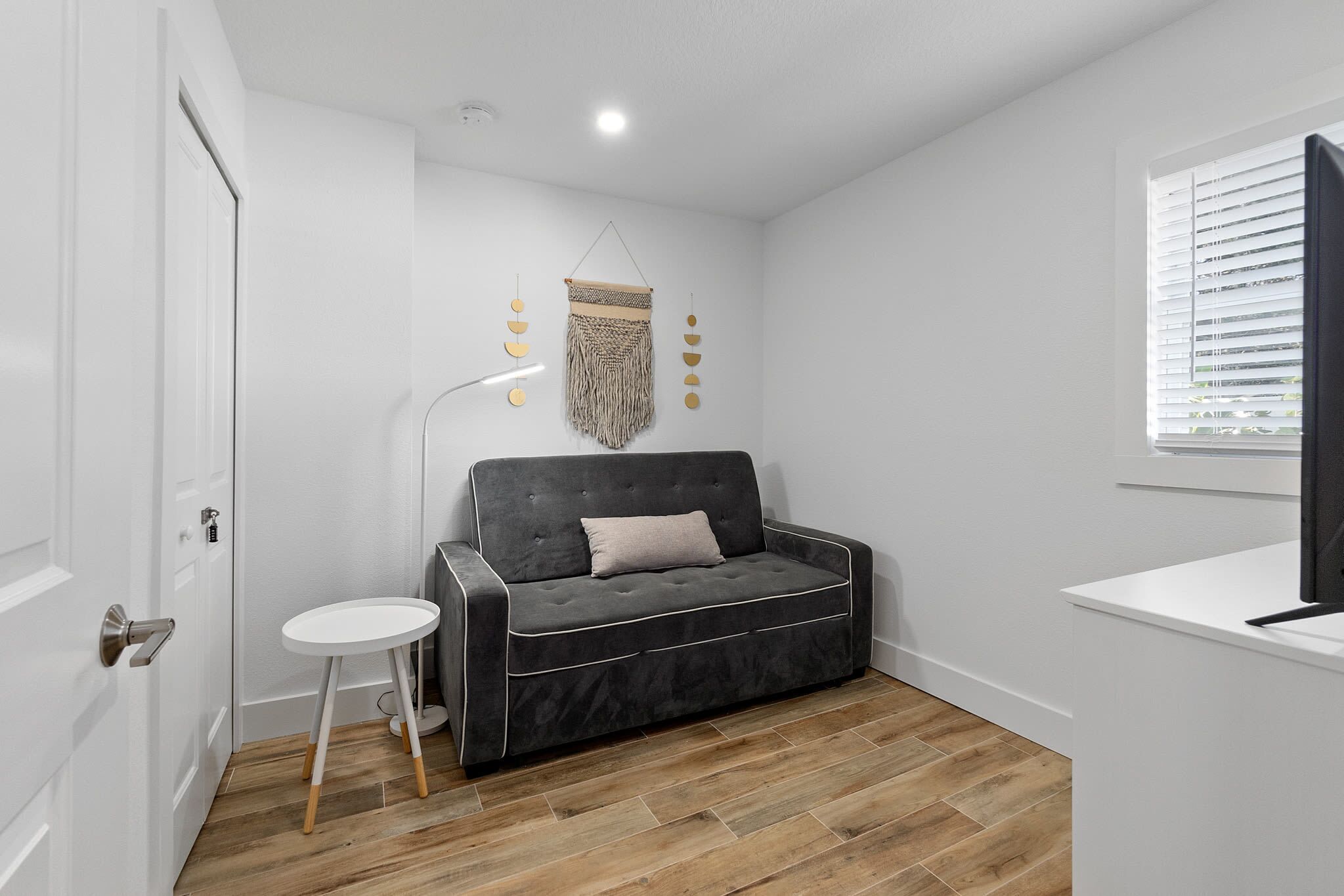 A room with a dark grey sofa, a small white side table, a wall-mounted TV, and a wooden floor.