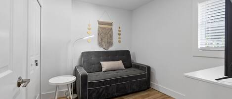A room with a dark grey sofa, a small white side table, a wall-mounted TV, and a wooden floor.