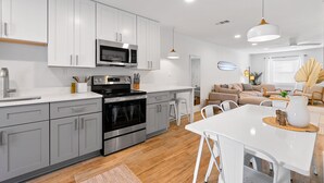 A modern kitchen with white cabinets, a stainless steel oven, and a microwave. There is a dining area with a white table and chairs.