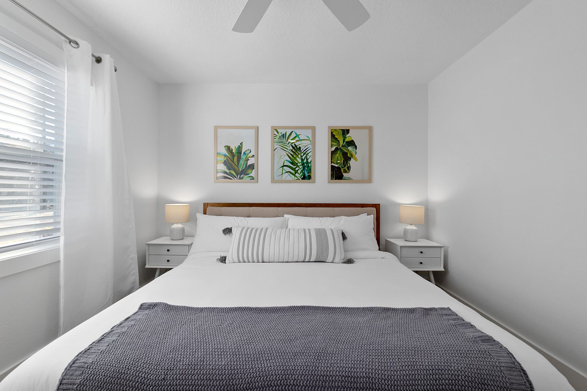 A neatly made bed with a gray blanket, two bedside lamps, and three framed botanical prints on the wall.