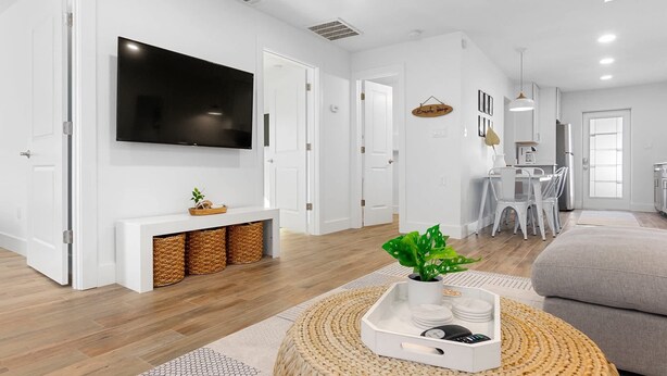 A modern living room with a flat-screen TV, a dining area with a table and chairs, and a kitchen with white cabinets and a refrigerator.