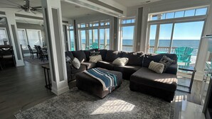 A spacious living room with a large sectional sofa, a dining area with chairs, and a view of the ocean through large windows. - beach front home pool  many new upgrades (North Topsail Beach)