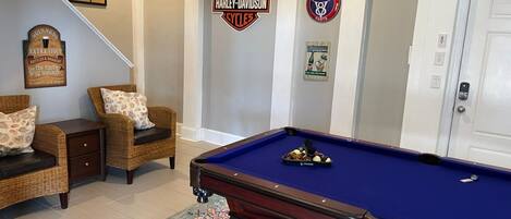 Una mesa de billar en una habitación con alfombra azul, dos sillas de mimbre y un cuadro en la pared.