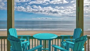 A wooden deck with turquoise Adirondack chairs and a small round table, overlooking a beach with a clear view of the ocean and a partly cloudy sky. - beach front home pool  many new upgrades (North Topsail Beach)