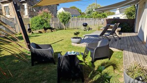 Een achtertuin met een houten terras, twee zwarte loungestoelen, een bijzettafeltje met bekers en een bloempot.
