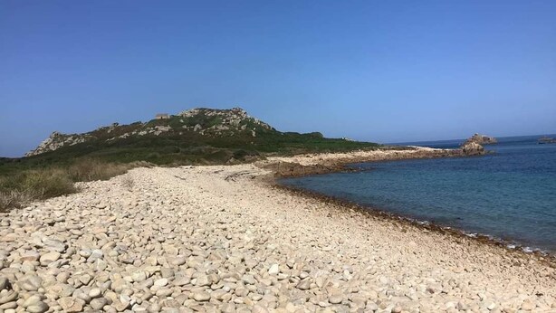 Een kiezelstrand met een rotsachtige heuvel op de achtergrond en een kalme zee.