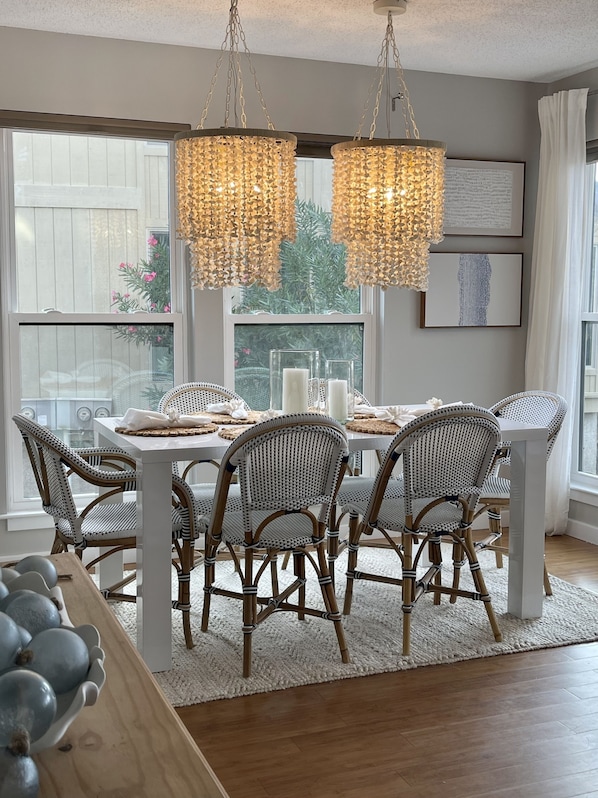 A dining area with a white table set for six, surrounded by wicker chairs, under a chandelier with hanging lights.